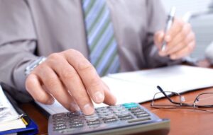 An accountant working on tax forms and filings with a calculator, papers and pens. There is a pair of eye glasses sitting on the table as well.