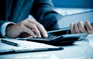 An accountant sitting at a desk working on a ledger with a calculator and iPad.