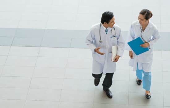 Two doctors walking across a room. One doctor is talking while the other is listening and carrying a blue folder.