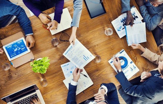 A collection of individuals sitting around a table exchanging ideas, designs, paperwork, screen shots and other collateral.