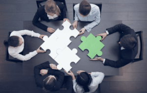 A group of six employees sitting around a table with 3 white puzzle pieces and a fourth green piece being put together as a group.