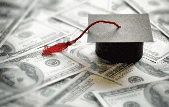 A stack of money piled on a desk with a graduation cap on top.