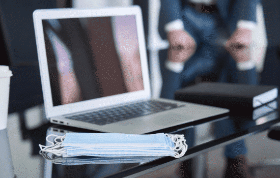 A laptop sitting on a glass desk with an KN-95 mask sitting beside it.