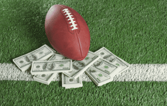 A stack of 100 dollar bills and a football laying against it on a dubber football field.