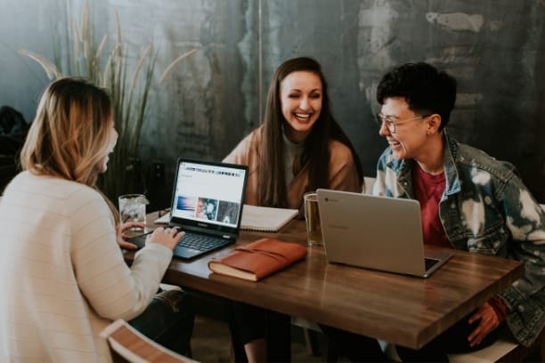 Three women sitting at a table laughing while working on laptops at a table.