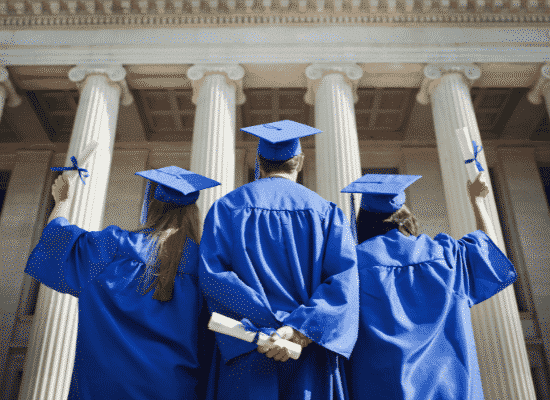 Three graduates holding diploma in front of a campus building all wearing blue cap and gown attire.