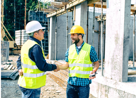 Two construction workers shake hands at a construction site. They are standing outside a partially constructed building with construction equipment around them.