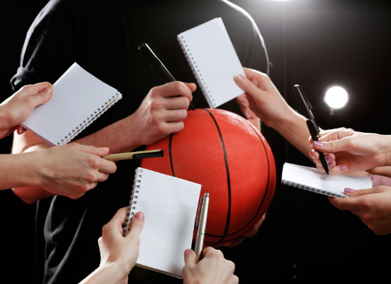 An athlete signs a basketball while fans hold out pens and pads of paper, waiting for their turn to get an autograph.