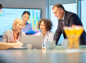 Two peole are sitting at a table looking at a laptop computer. A man and a woman are standing behind them looking at the same screen. There is a screen in the background that is displaying a chart. Coffee mugs and pencils are on the table in front of the people.