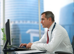 A physician sits at his desk looking at a computer and appears focused on the computer screen. Highrise buildings are visible through his office window.