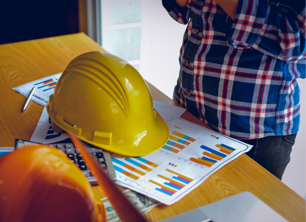 Two hard hats are on a desk in an office next to a calculator and financial reports. A person is standing over the desk.
