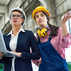 Two women wearing hard hates stand next to each other in a in a ware house. One is holding a laptop and dressed in a business suit. The other is wearing an apron and hearing protection around her neck.