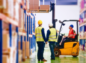 Two workers wearing hard hats are standing in a supply warehouse watching a third worker on a forklift loading large boxes onto the shelves.