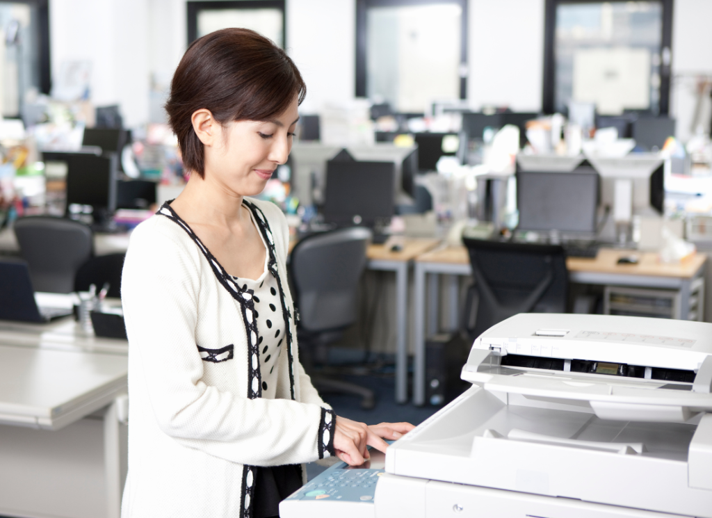 A woman is using a copy machine in an office setting. She is facing the copier while smiling and pressing a button.