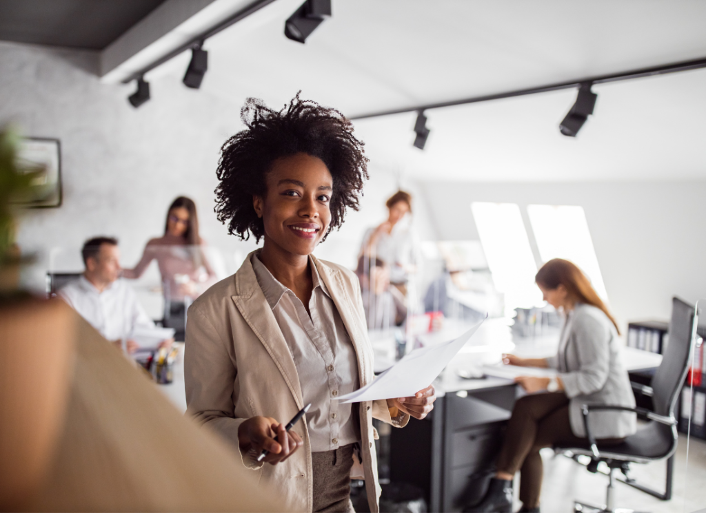 People are working in an office setting. The image focuses on a woman holding a pen and paper and smiling.