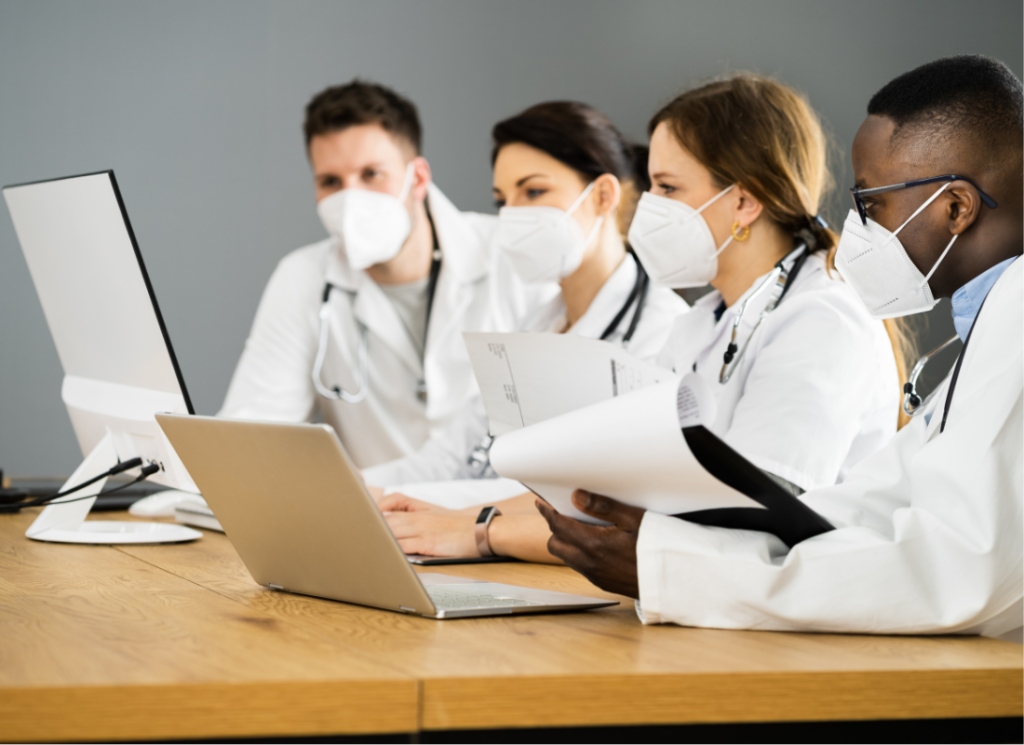 Four physicians are sitting at a table looking at a computer screen and a laptop. All four physicians are wearing a mask and one is holding a clipboard and papers..