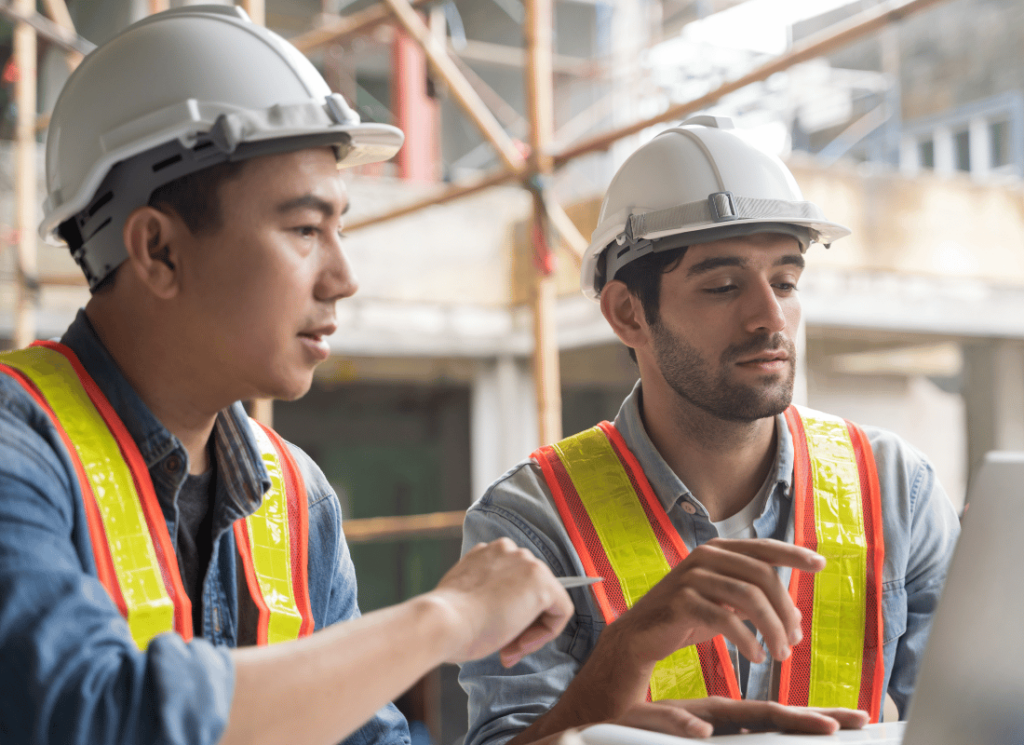 Two construction foremen reviewing plans on a laptop on a job site.