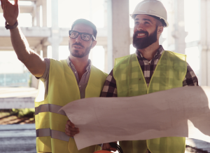 Two construction workers are standing together at at a construction site and both are wearing yellow vests. One worker is pointing to something off camera. The other worker is wearing a hard hat and holding blueprints.