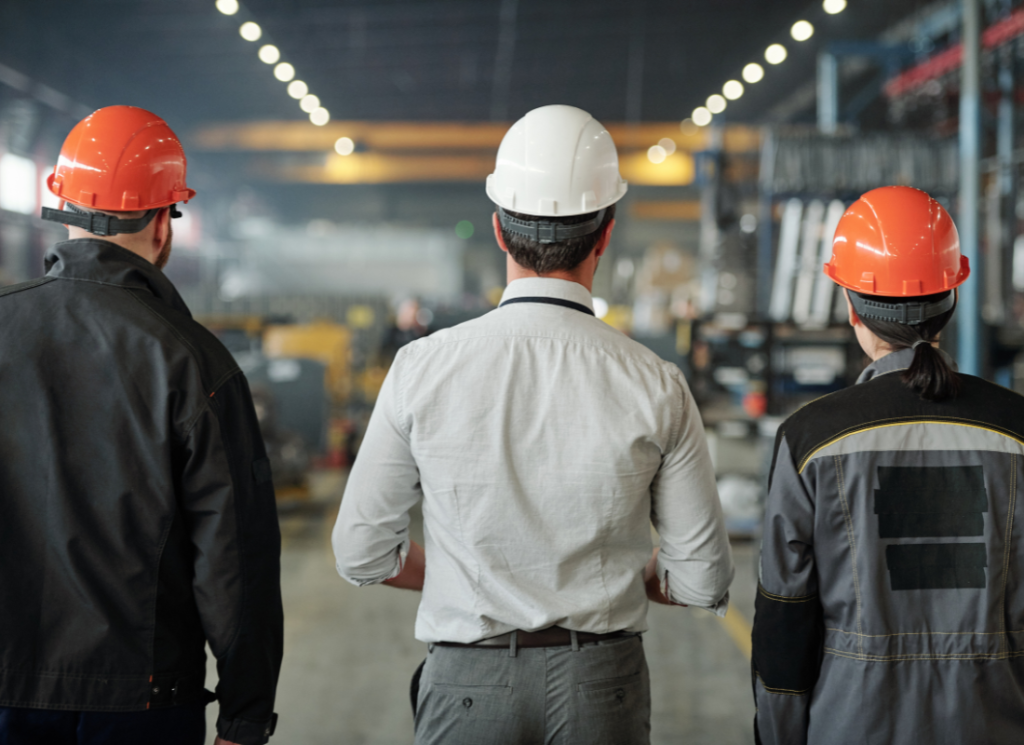 The backs of three workers are standing in a manufacturing warehouse. There is manufacturing equipment in the background.