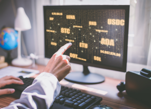 Someone is pointing to a computer monitor on a desk. The monitor is displaying various cryptocurrency names.