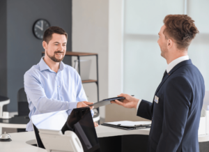 One person is standing behind a front desk and handing a folder to the person in front of the desk.