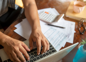 A laptop computer is shown on a desk with tax documents, a calculator, and a pen. There is a person sitting at the desk and typing on the laptop. Only the person's arms and hands are shown.