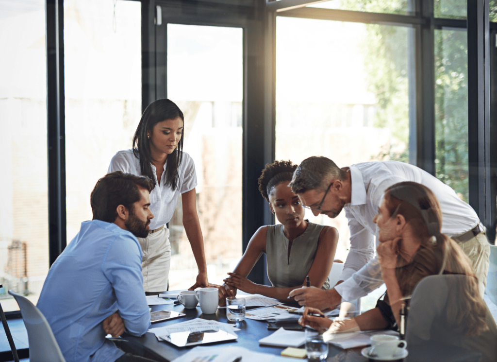 5 people are gathered around a table ion an office building. One of them is writing on paper while the others look on. There are papers and coffee mugs on the table and a large window in the background.