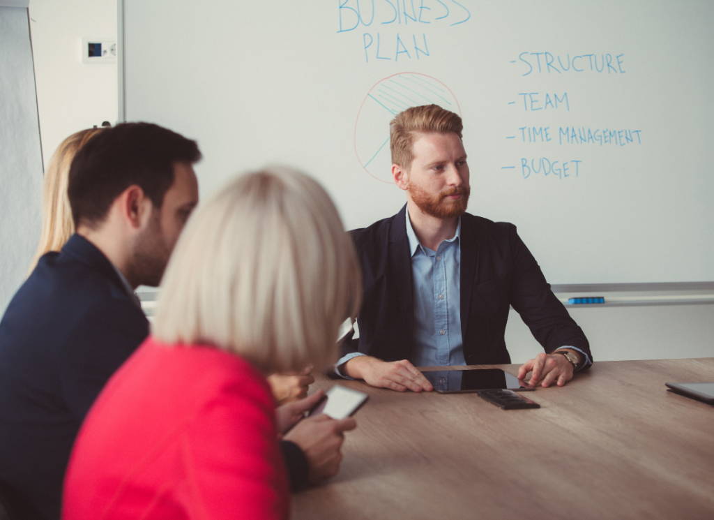 People are sitting at a table in a boardroom. Three people are shown in the photo and there is a white board with writing in the background.