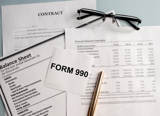 Finance related documents are laying on a table with other papers. One piece of paper says "Form 990". One of the papers says "Balance Sheet" and the bottom paper says "Contract". There is a pen and a pair of glasses on top of the papers.