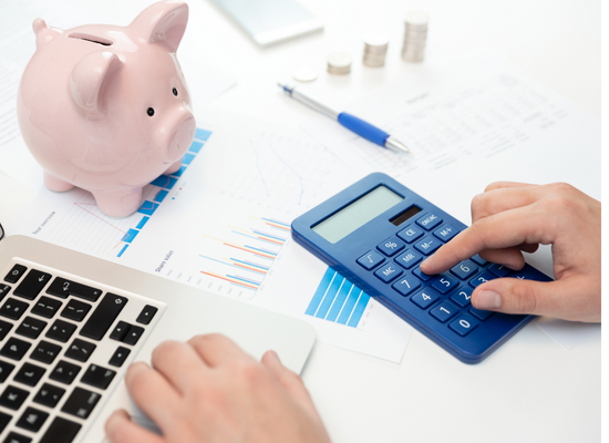 Two hands are shown typing on a calculator and a laptop keyboard on top of a table. A piggy bank, pen, paper, and three stacks of coins are also shown on the table.