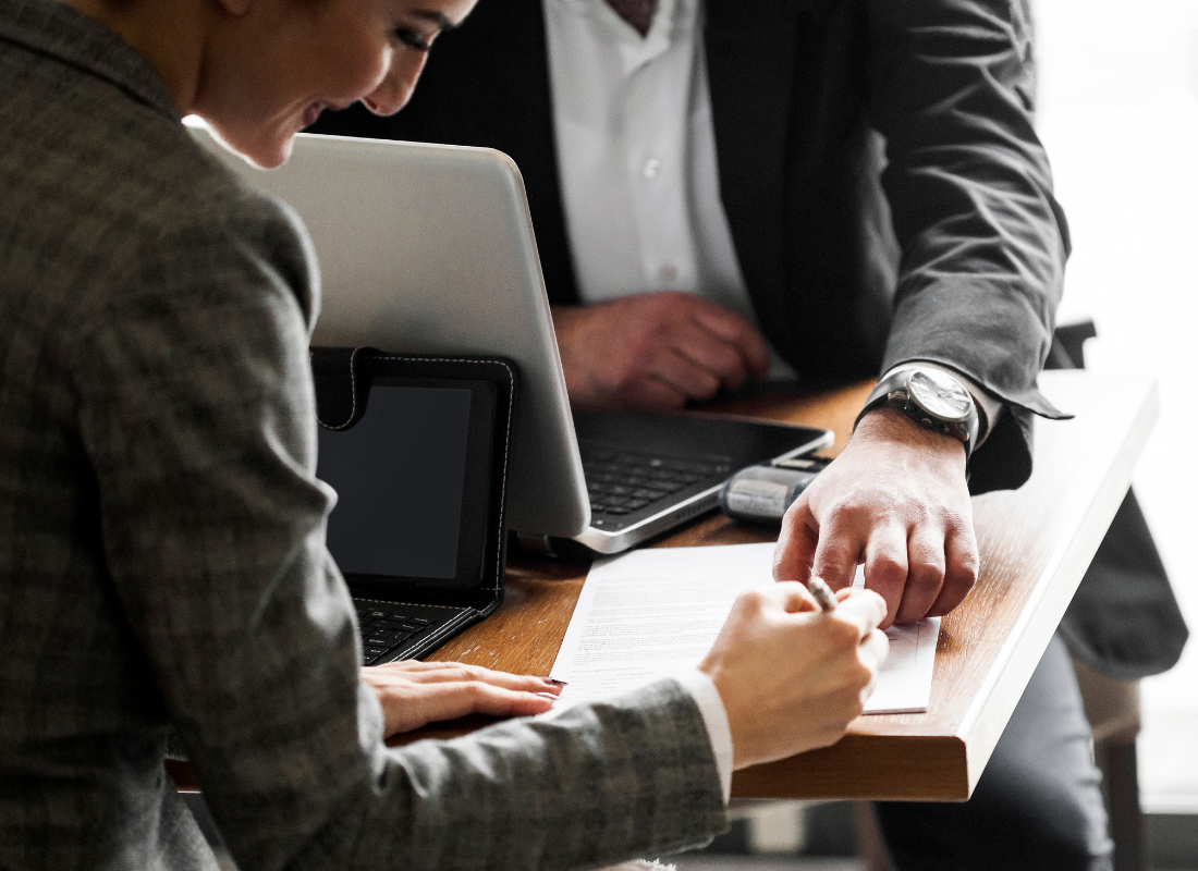 A business person is signing a paper and sitting across from someone else in a business suit. Both of them have laptop computers open. The person across the table is pointing at the paper.