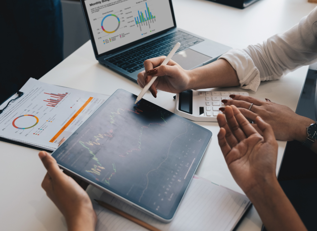 A computer, tablet, calculator, and graphs are on a desk. People are examining the blockchain charts shown on the tablet.