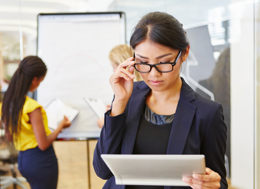 A business woman wearing a suit and glasses is reading a sheet of paper while two others our assessing a white board in the back ground.