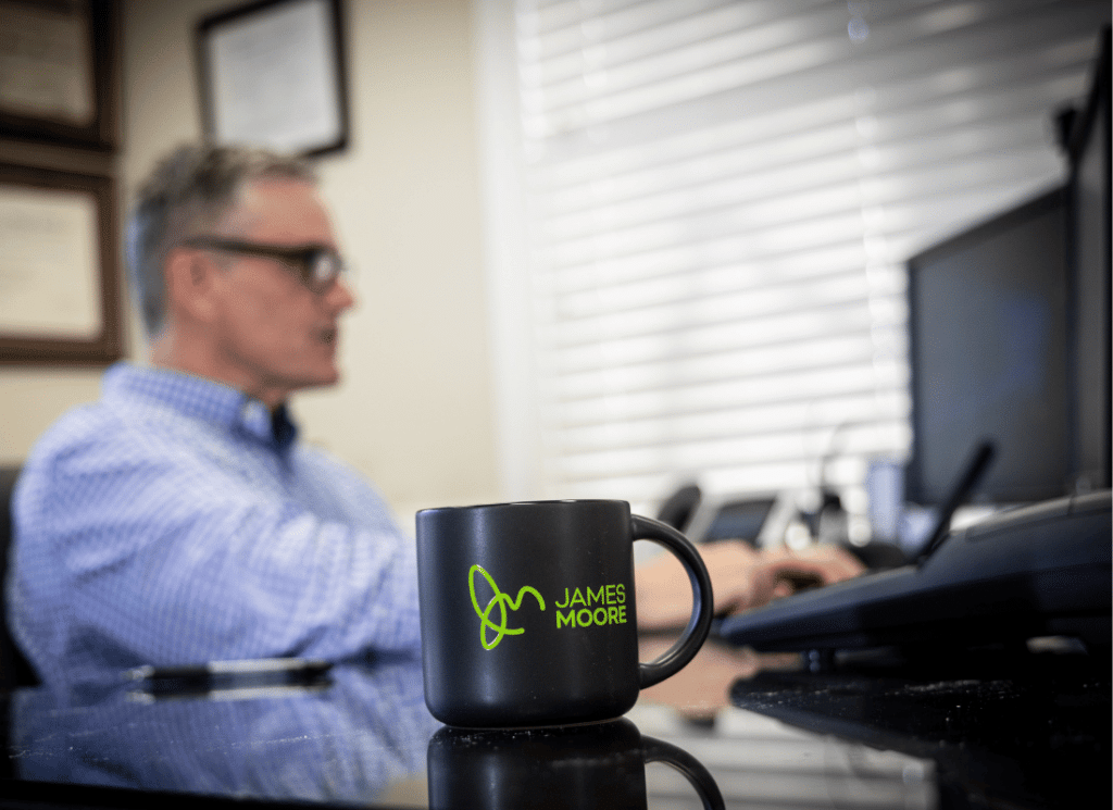 A coffee mug with the James Moore & Co logo sits on a desk in front of a man who is working on his computer.