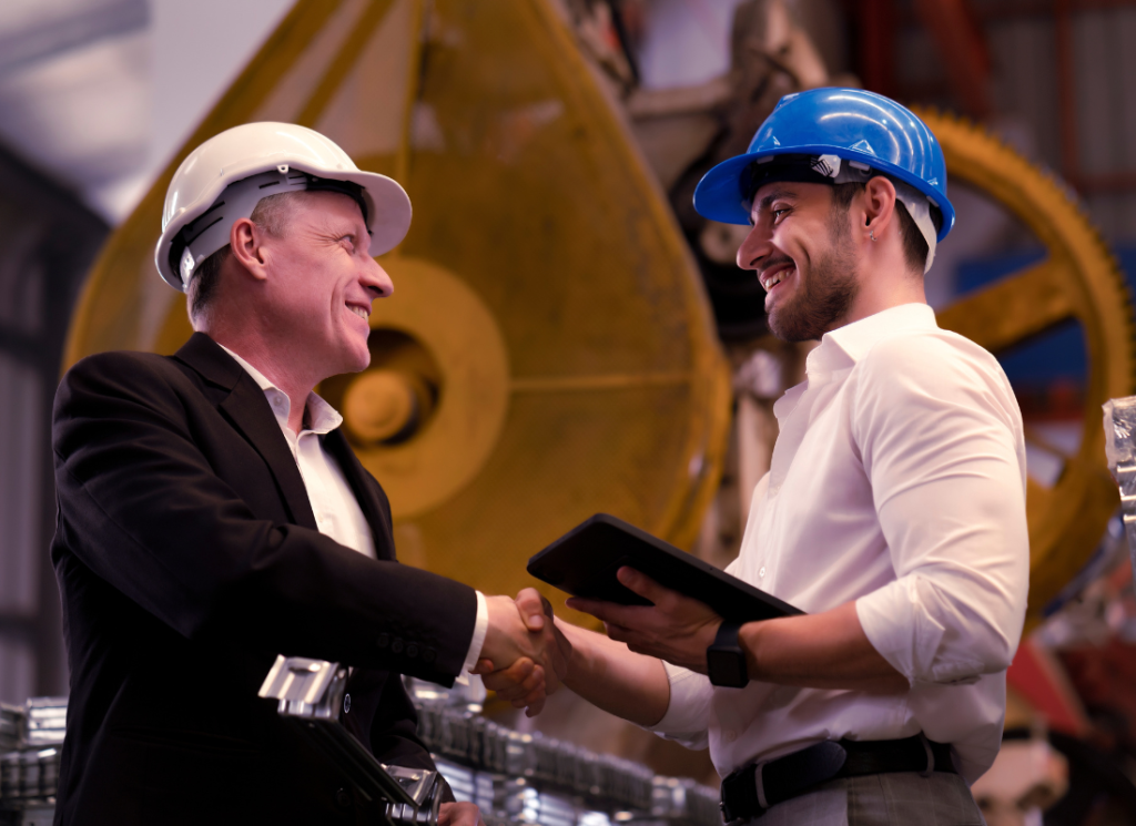 a business owner and employee smile and shake hands in a manufacturing facility. Both are wearing hard hats and there is machine equipment in the background.