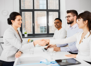 A business person is sitting across a from three employees at a table in an office setting. The business person is shaking hands with one of the employees.