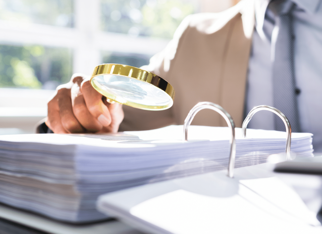 An auditor reviews documents in a binder with a magnifying glass.