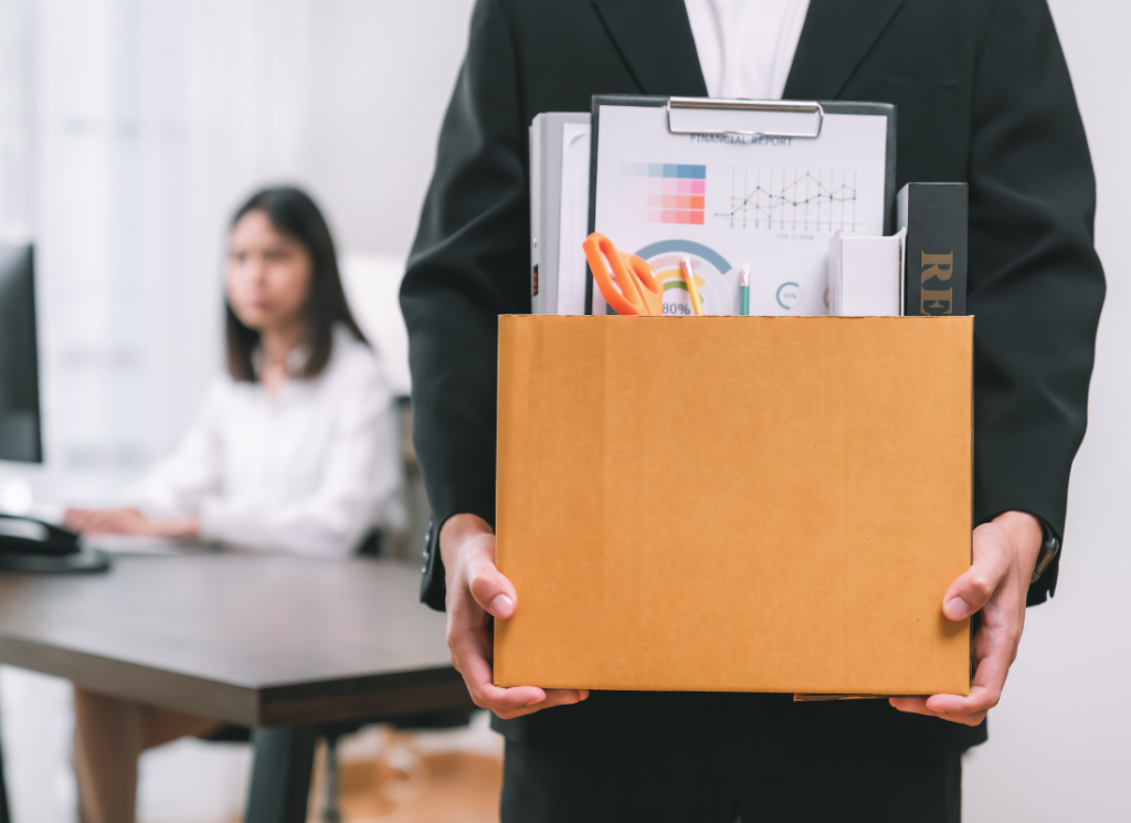 An employee is in an office holding a box of office supplies and personal items. In the background, a person is at a desk on a computer.