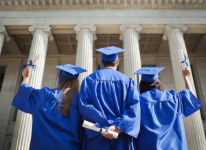 Three college graduates, wearing a cap and gown, stand outside a building on a university campus.