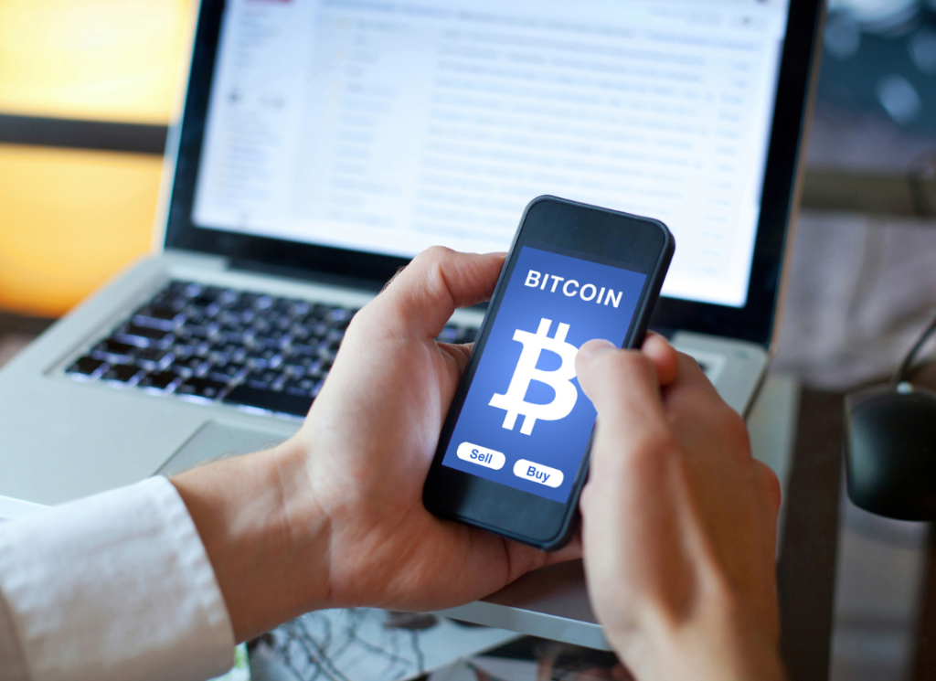 A smartphone is pictured with a cryptocurrency logo displayed on the screen. A laptop computer is open in the background.