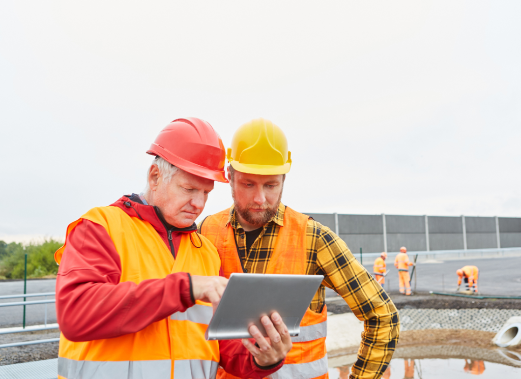 Two construction workers are standing at a construction site, wearing hard hats and vests, while other workers are in the background. One of them is holding a laptop and both are looking down at the screen.