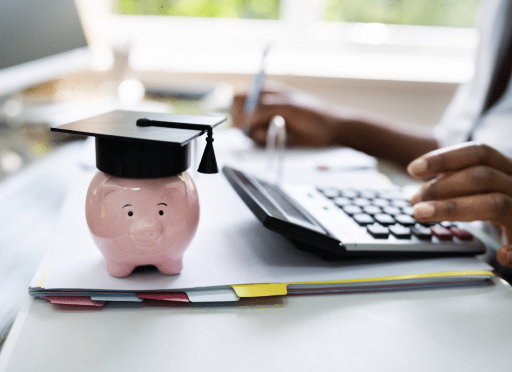 An accountant is at a desk typing in a calculator and writing with their other hand. there is a piggy bank wearing a graduation cap on the desk.