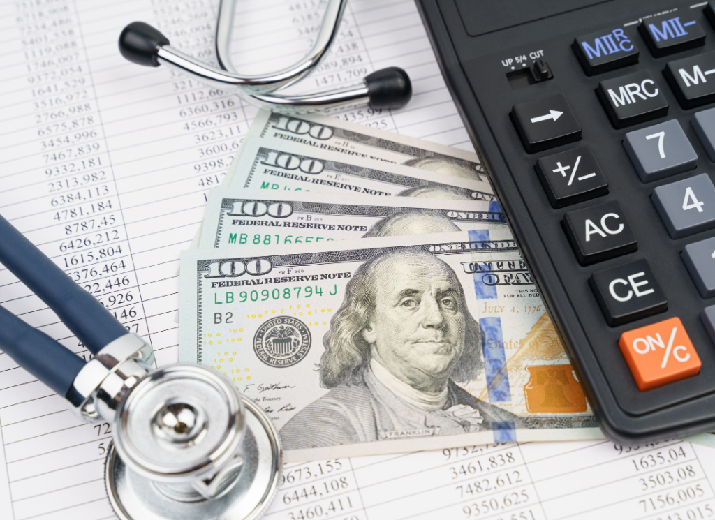 A stethoscope, a calculator, and cash in the form of one hundred dollar bills are pictured on top of financial documents.