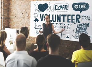 A person is speaking in front of a group of people. She is standing at the front of a crowd and pointing toward a banner on the wall that says 