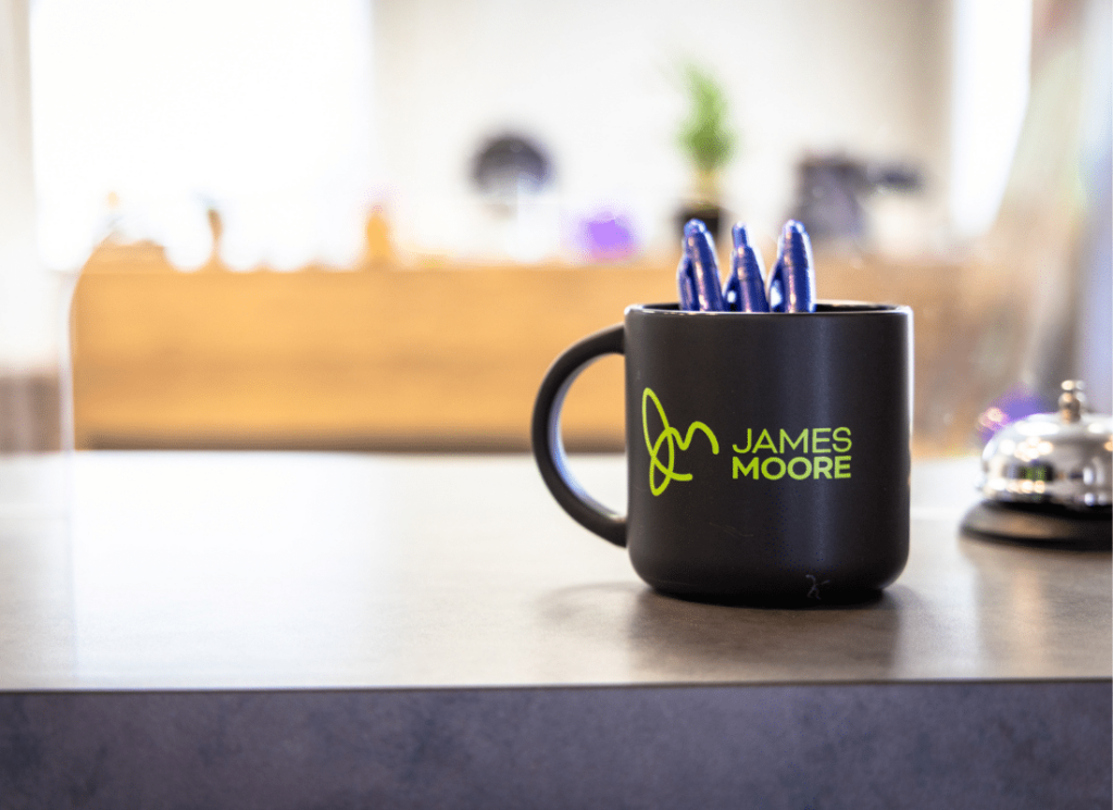 A coffee mug with the James Moore & Co. logo, and three blue pens inside the mug, on a desk with an office setting in the background.
