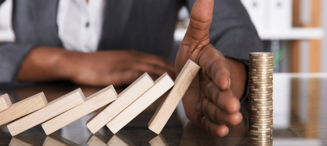 Close-up Of A Human Hand Stopping Wooden Blocks From Falling On Stacked Coins Over Desk.