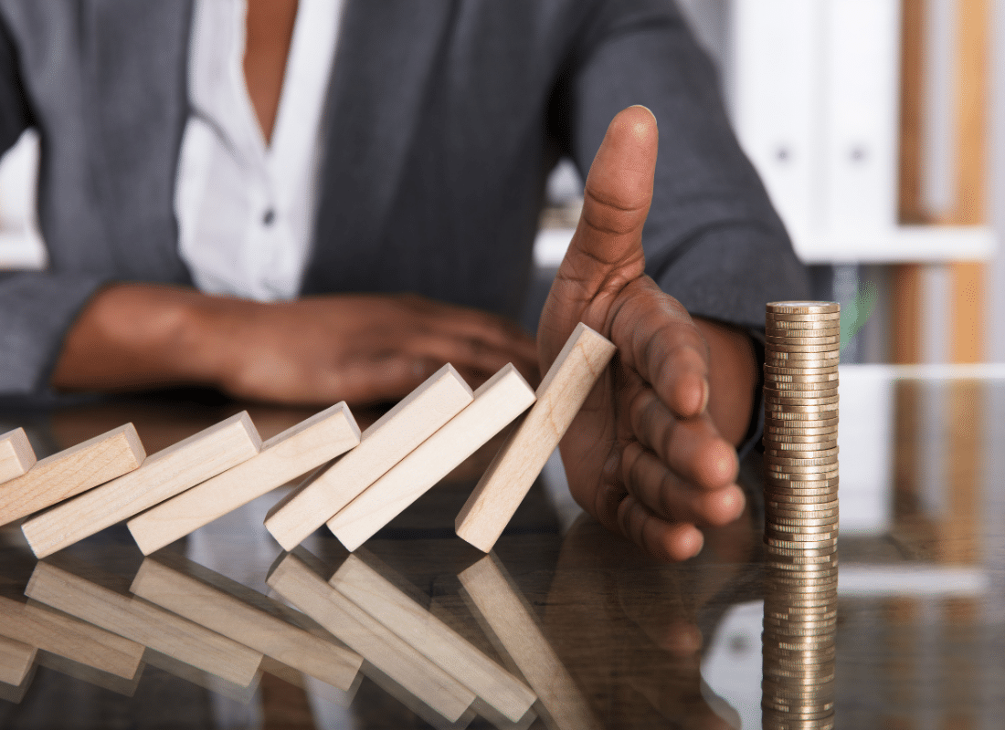 Close-up Of A Human Hand Stopping Wooden Blocks From Falling On Stacked Coins Over Desk.