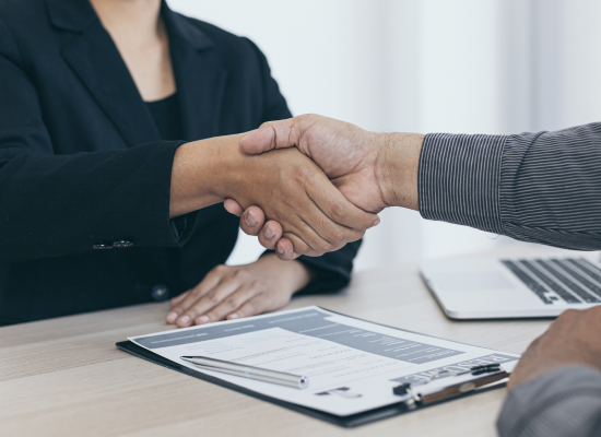 Two people are sitting at a desk for an interview and are shaking shaking hands across the desk. There is a computer and resume on the desk.