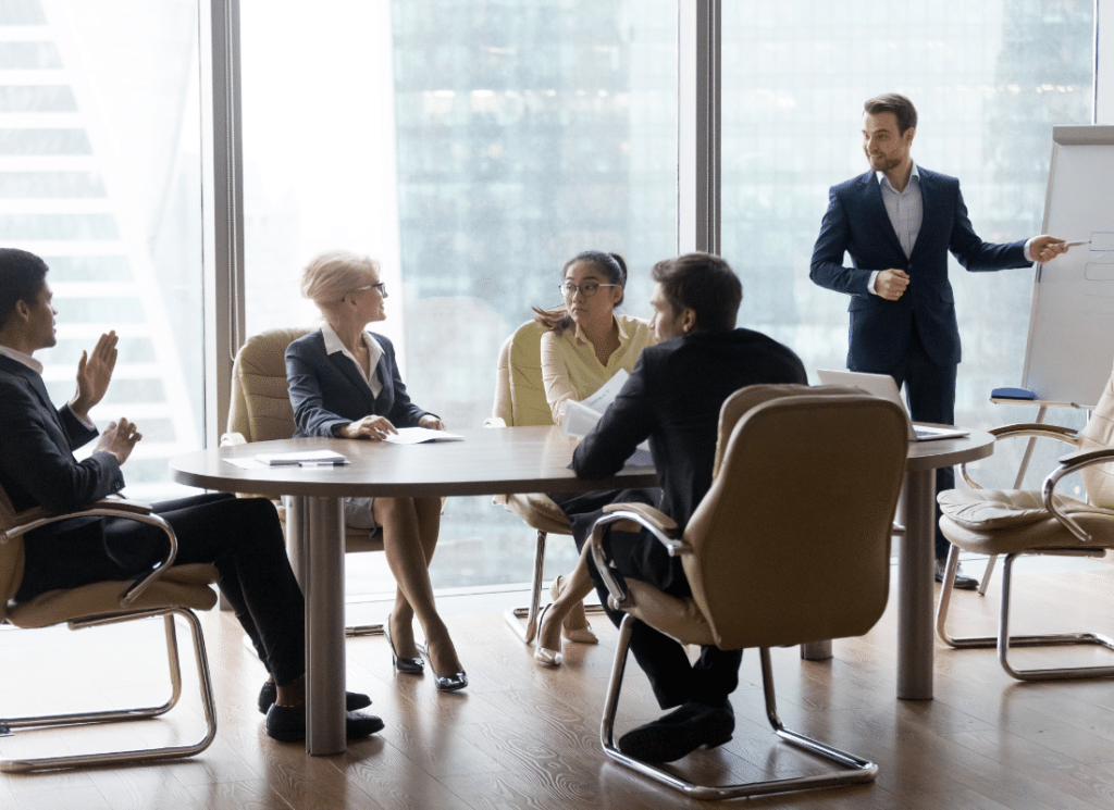 A group of five people are in a business meeting. The person leading the group is standing in front of a whiteboard, while the rest of the group is seated at an oval table.