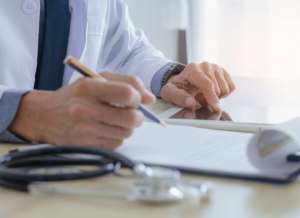 A doctor sitting at a wooden desk in a hospital or clinic. The doctor is working on a tablet computer and reading a document on a clipboard. He is holding a pen and there is a stethoscope on the wooden desk.